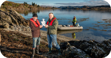 One of them is holding a white object, which appears to be a small model or sculpture, and both are smiling. Behind them, a small inflatable boat with a person standing inside is anchored near the shore. The background features a calm lake with mountains and a partly cloudy sky, creating a scenic, outdoor setting.